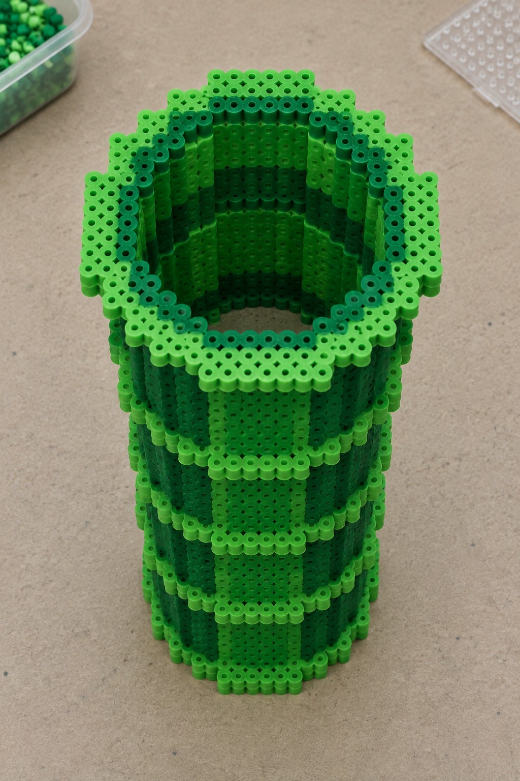 Tall 3D Perler bead pipe made from stacked layers of green beads in several shades, forming a hollow square-top cylinder.