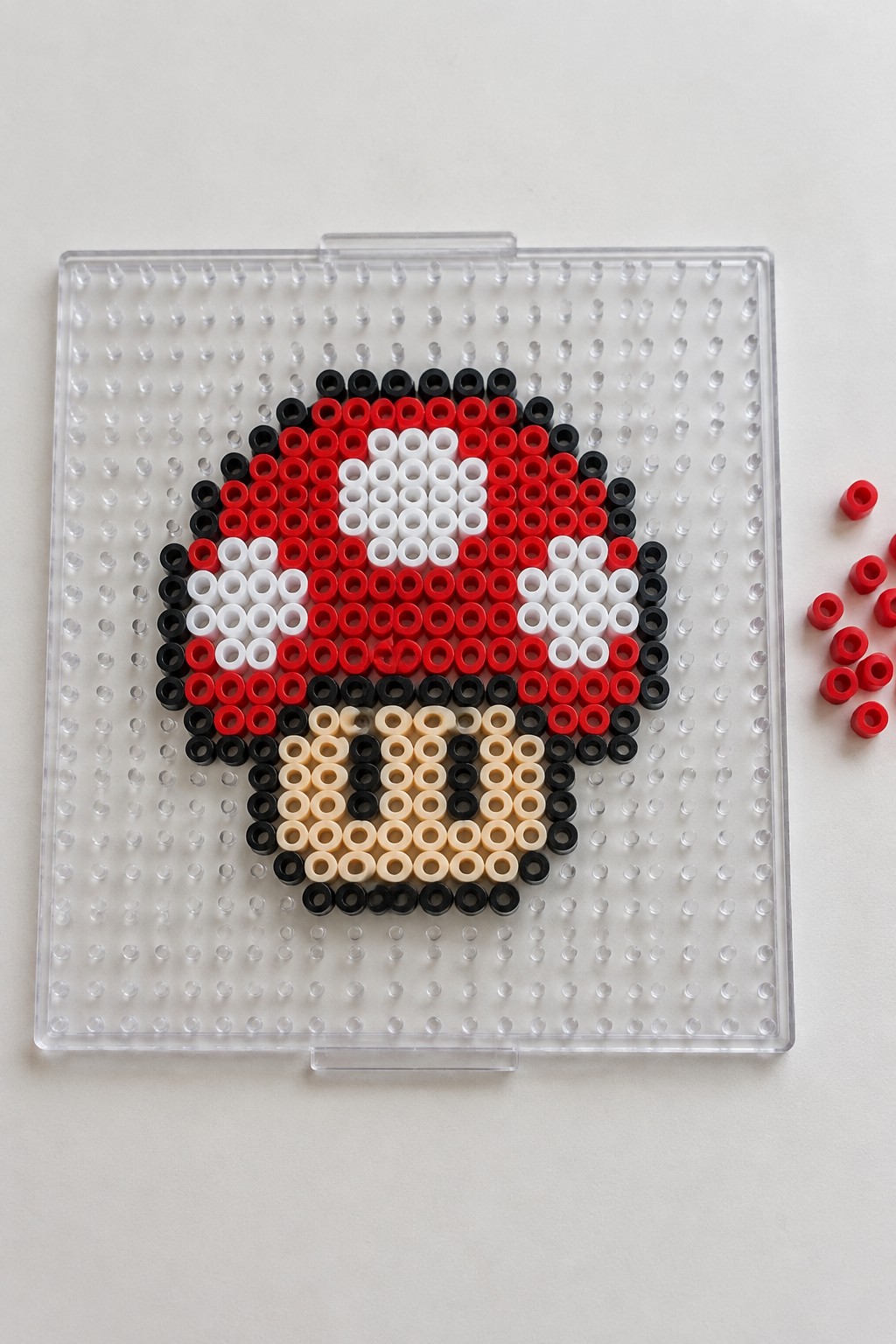Perler beads on clear pegboard form red white black beige circular pattern with loose red beads