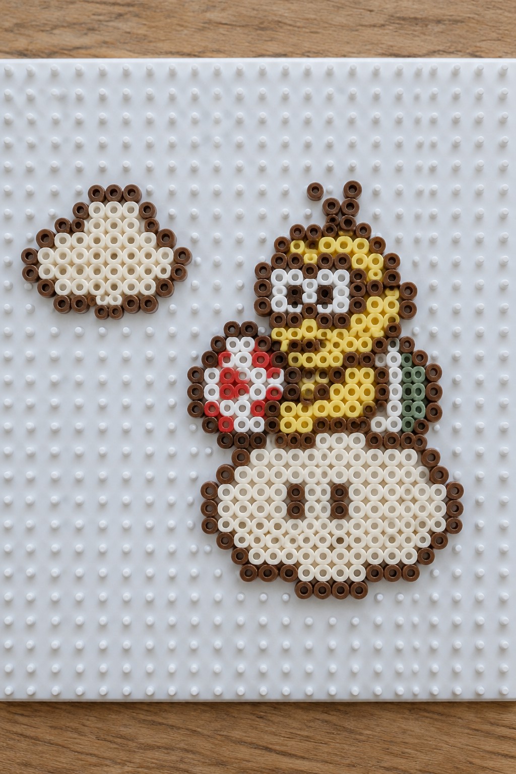 Perler beads forming a brown-outlined cartoon figure with yellow face beside small circular design on pegboard
