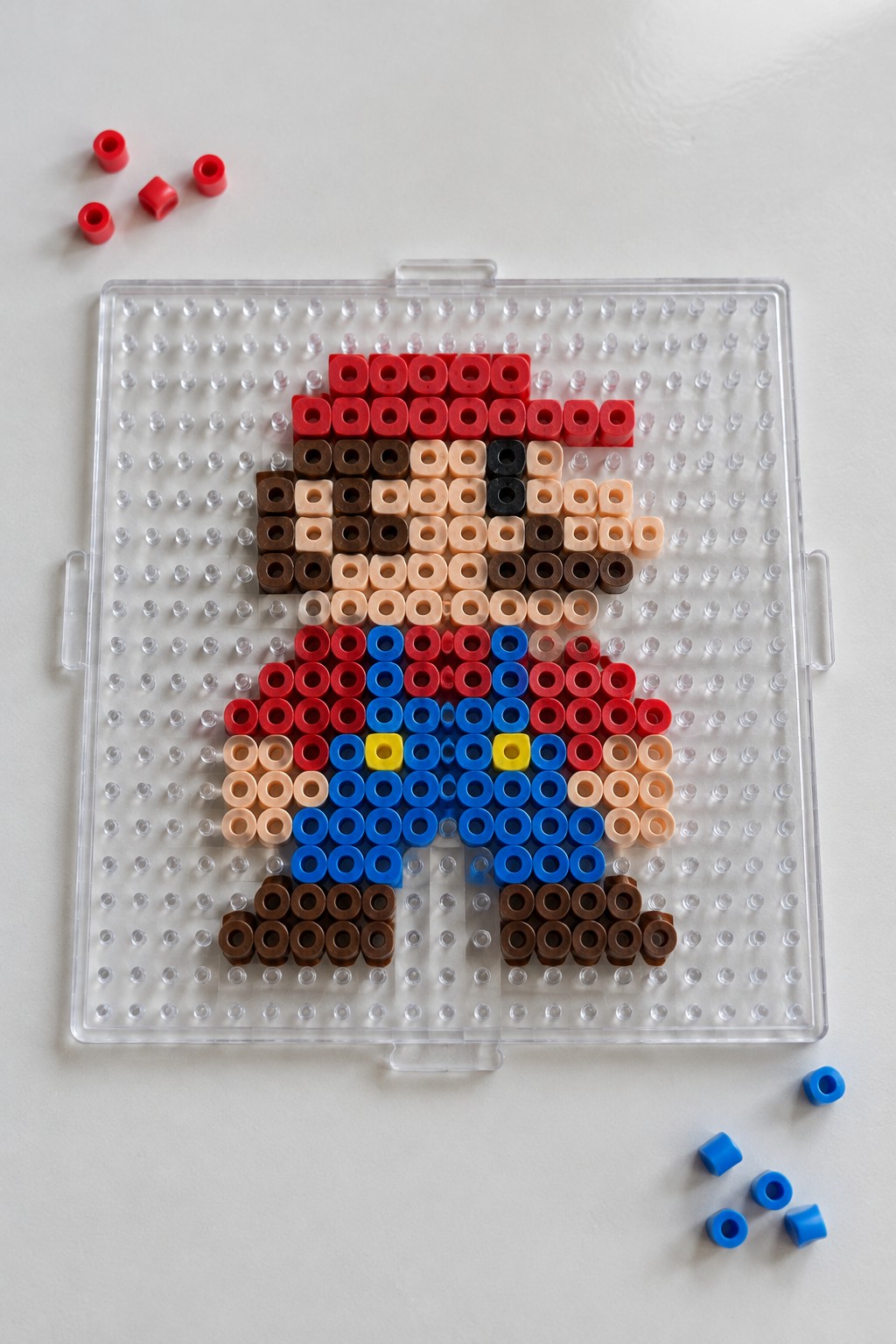 Perler beads forming Mario in red hat and blue overalls on clear pegboard with loose beads