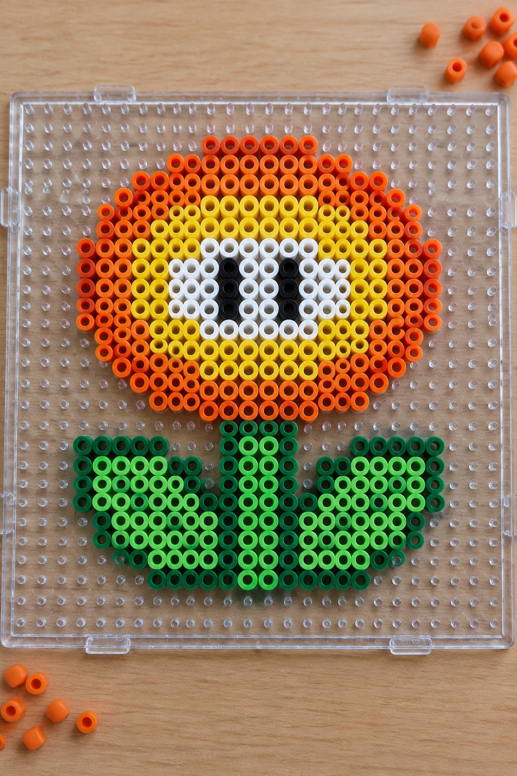 Perler bead flower with orange petals, yellow center, green leaves on pegboard.