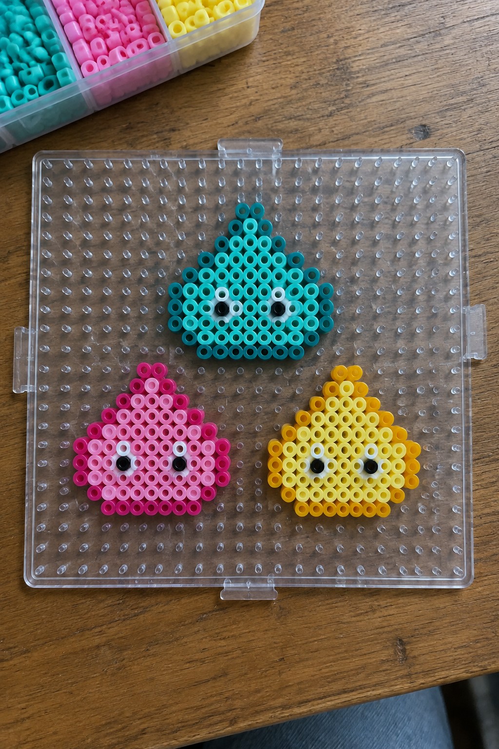 Three colorful teardrop perler bead figures with faces on clear pegboard beside bead tray