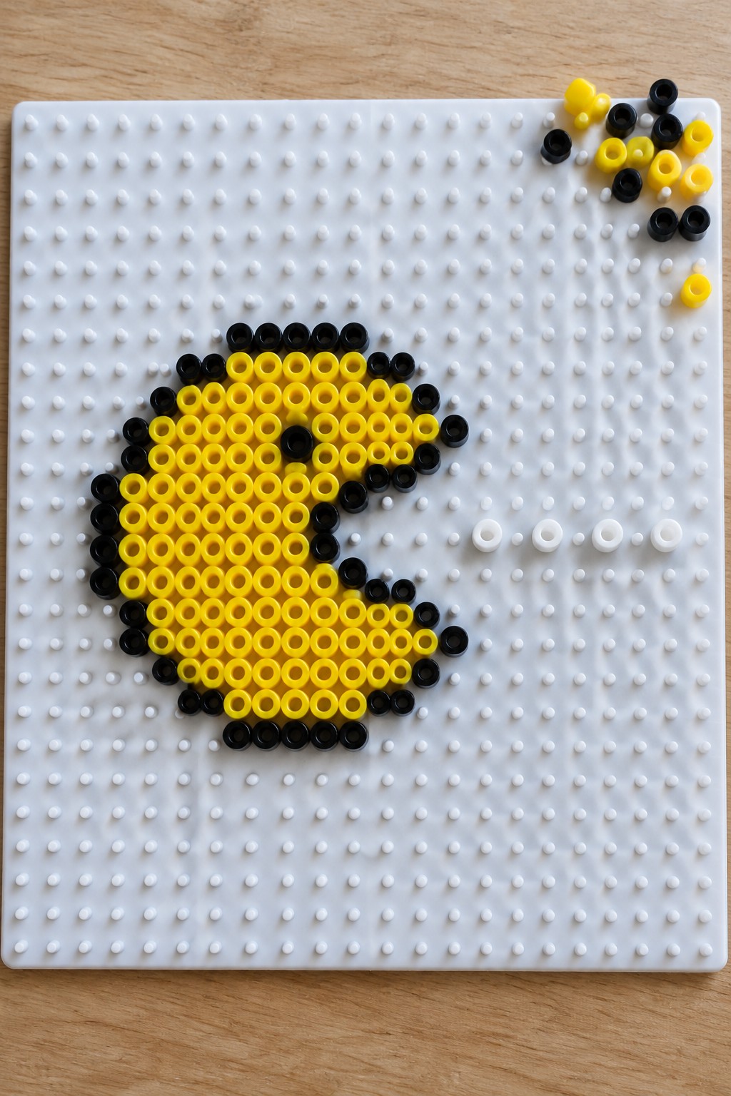 Yellow and black Perler beads forming Pac-Man on a white pegboard with loose beads nearby.