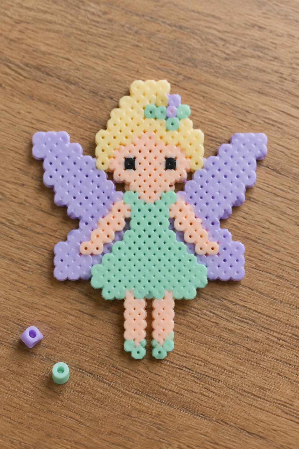 Perler bead fairy with yellow hair, mint green dress, and purple wings on wood