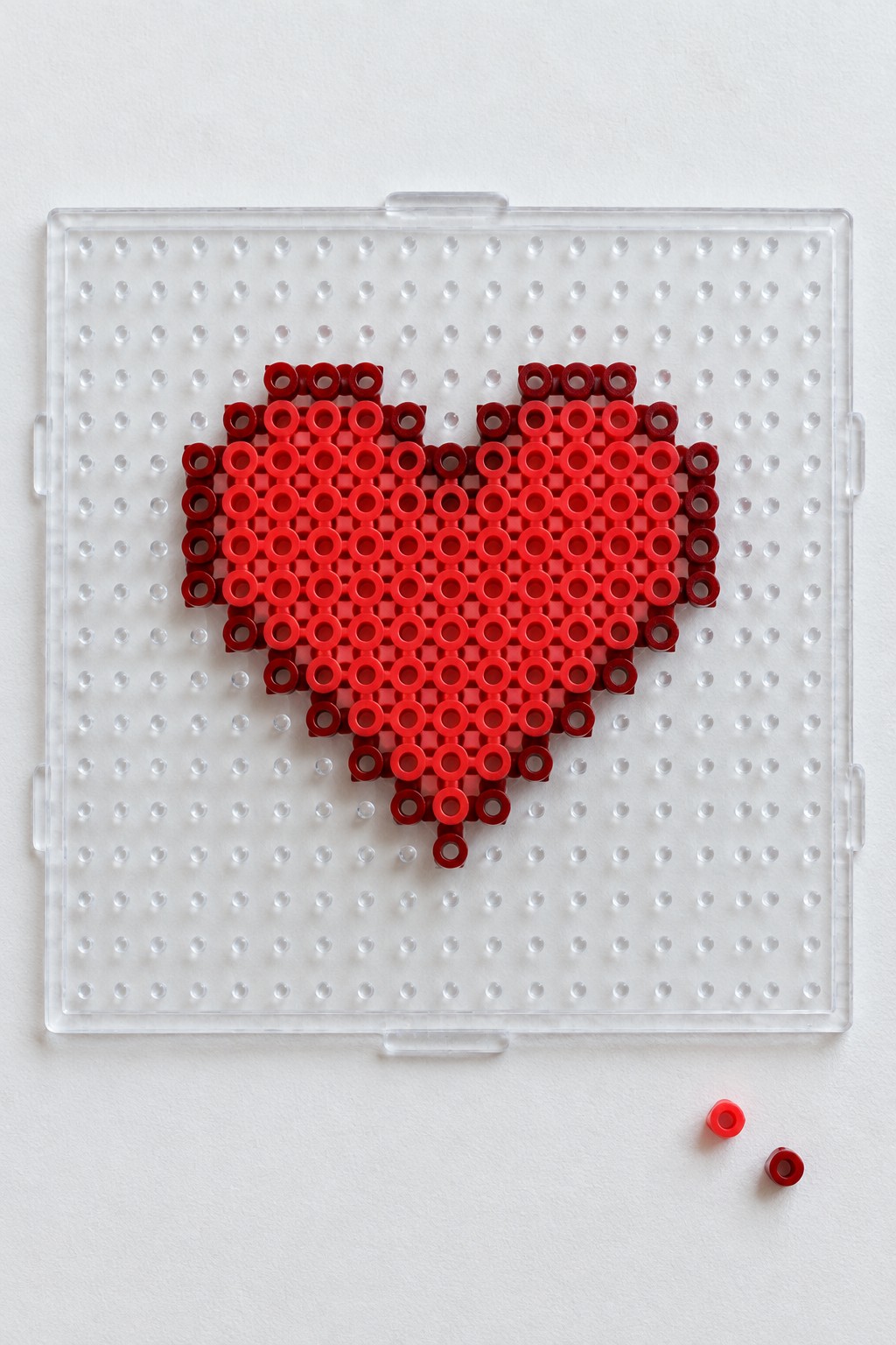 Red pixelated heart of beads on clear pegboard with two loose beads nearby.