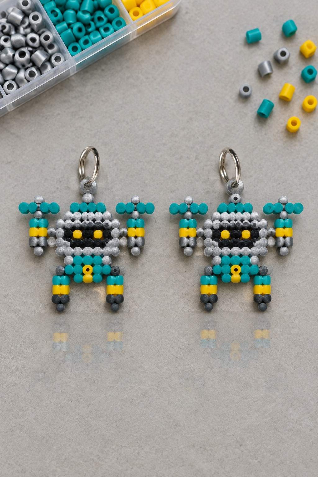 Two perler bead robot keychains in teal, yellow, and gray beside bead organizer