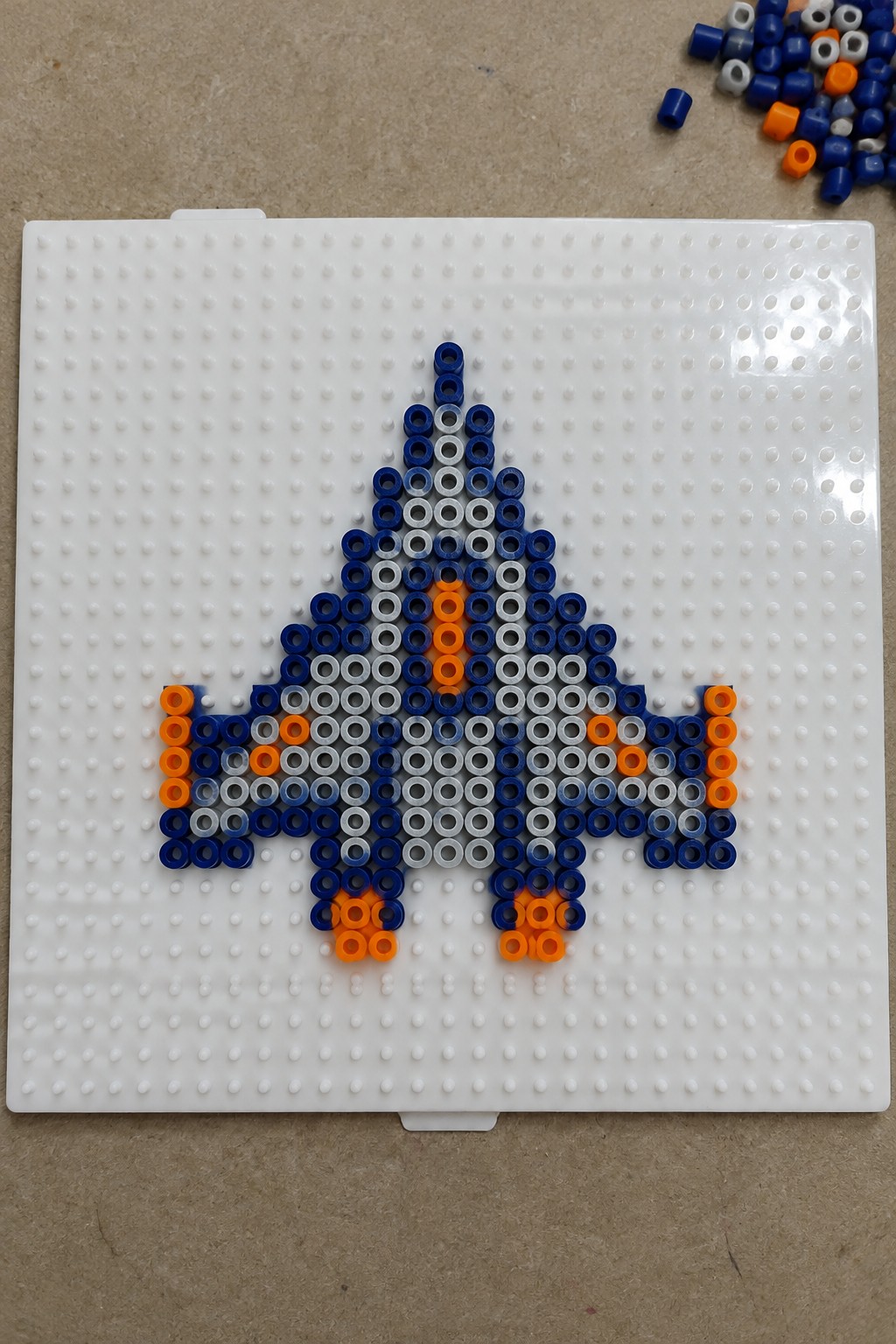 Perler beads forming a blue jet with orange accents on white pegboard